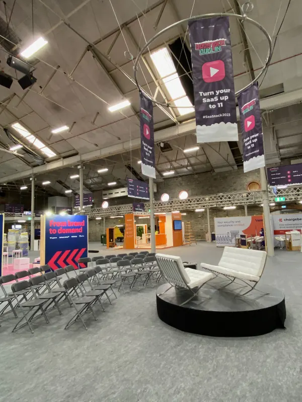 Behind the scenes at SaaStock Europe 2023 — empty Podcast Stage seating area with white Barcelona chairs on a round platform and 'Turn your SaaS up to 11' banners overhead, Dublin