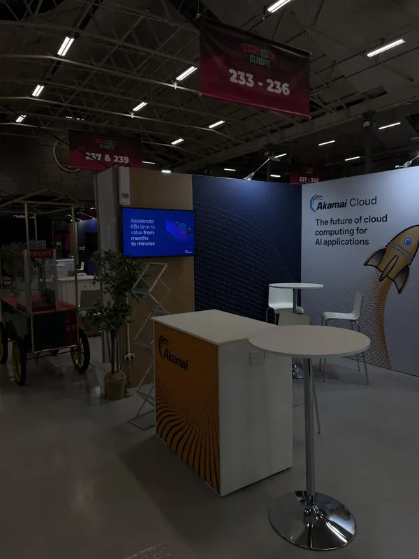 Before doors at SaaStock Europe 2025 — Akamai Cloud booth at 233–236 mid-setup with low light, 'The future of cloud computing for AI applications' wall just lighting up