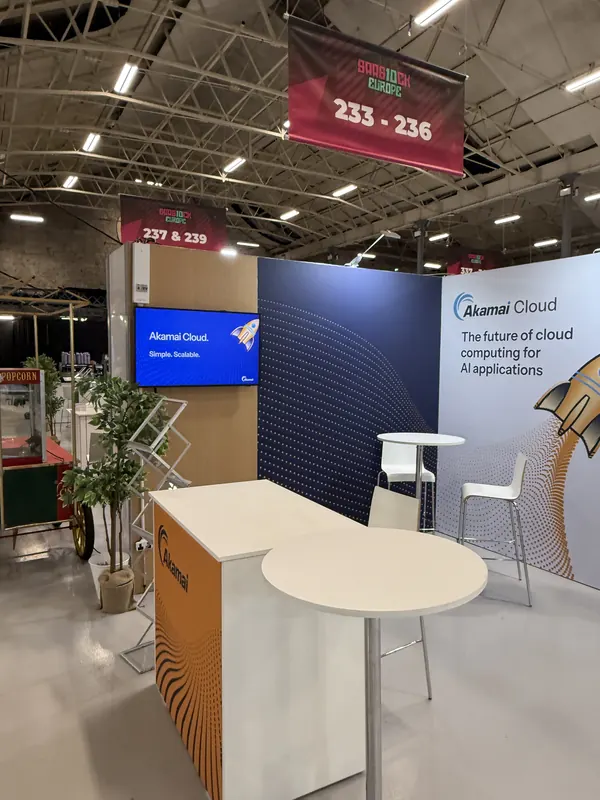 Before doors at SaaStock Europe 2025 — Akamai Cloud booth fully lit with the 'The future of cloud computing for AI applications' message and the orange rocket-burst illustration on the meeting counter