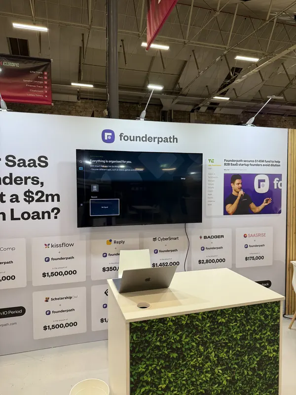 Before doors at SaaStock Europe 2025 — Founderpath booth pre-event with the 'Dear SaaS Founders, Want a $2m Term Loan?' wall, founder-deal grid (Kissflow $1.5M, CubeSmart $350K, Badger $2.8M, etc.) and live-demo monitor