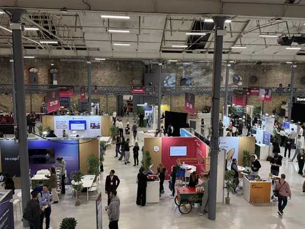Live during SaaStock Europe 2025 — wider aerial of the expo hall from the balcony showing booths, networking lounges, and attendee foot traffic between sessions