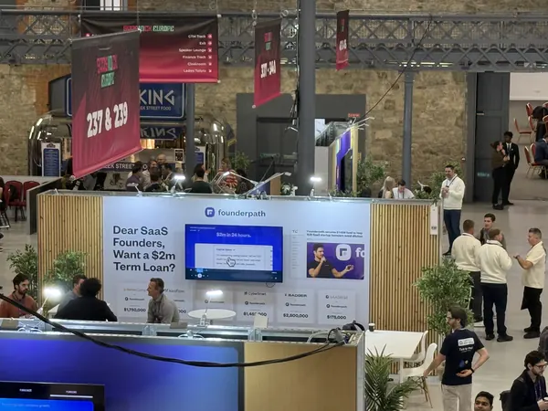 Live during SaaStock Europe 2025 — Founderpath sponsor booth viewed from the balcony, 'Dear SaaS Founders, Want a $2m Term Loan?' wall facing the aisle, attendees walking past