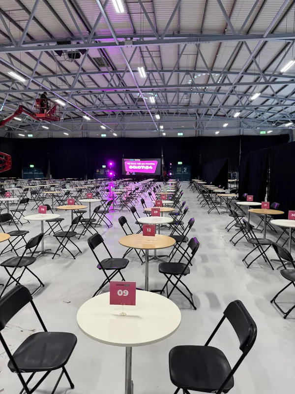 Behind the scenes at SaaStock Europe 2025 — wide-angle view of the empty 1:1 meeting floor showing rows of numbered tables stretching toward the meetup stage and pink uplighting