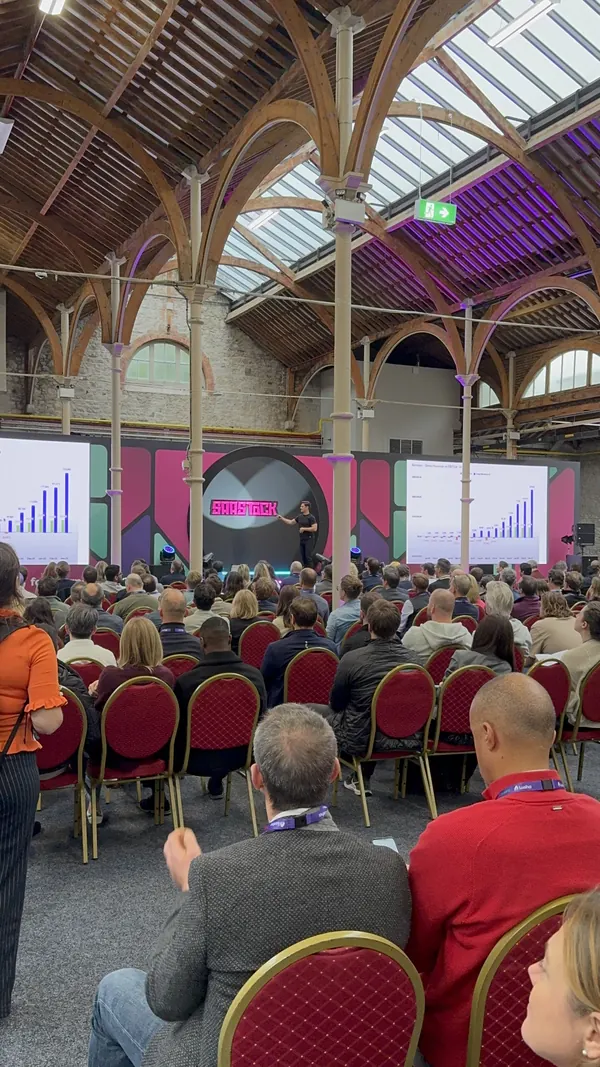 Live during SaaStock Europe 2025 — from-row view of the SaaStock mainstage with twin growth-chart slides flanking the SaaStock backdrop, audience seated in front