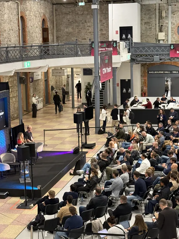 Live during SaaStock Europe 2025 — close-up of a speaker on the GTM Track stage with a slide reading '+1,200,000' (likely an ARR milestone), Victorian clock and ironwork in the venue above