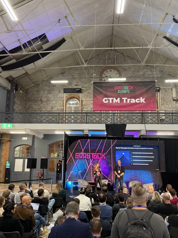 Live during SaaStock Europe 2025 — GTM Track stage shot showing two operators in conversation, audience seated, the GTM Track banner overhead and the SaaStock arch backdrop