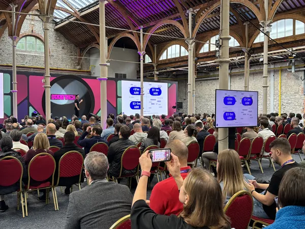 Live during SaaStock Europe 2025 — SaaStock signature arch backdrop on the mainstage with a seated audience in front, glass roof and wrought-iron columns of the RDS visible above
