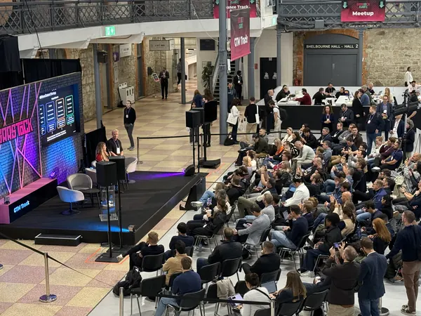 Live during SaaStock Europe 2025 — side angle showing a speaker on stage, full audience on the right side of the frame, GTM Track signage above on the mezzanine