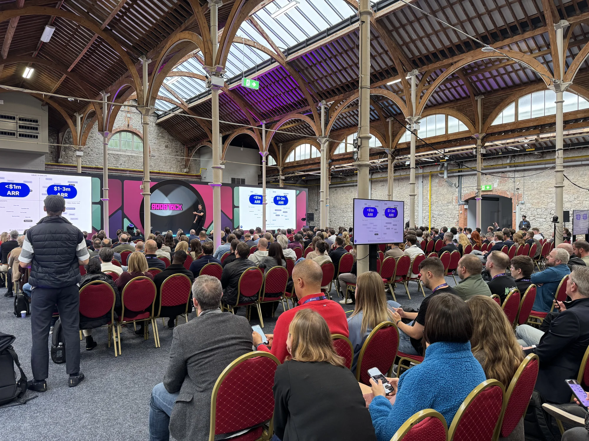 Mainstage at SaaStock Europe 2025 under the Victorian iron-and-glass roof of the RDS in Dublin — a packed audience of B2B SaaS founders watching the opening keynote on the GTM Track