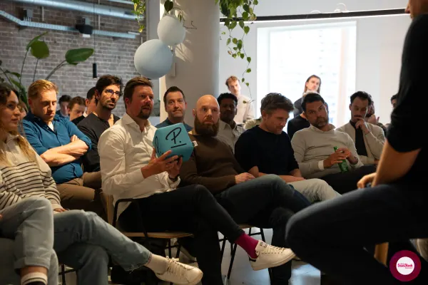 Live at SaaStock Local Amsterdam Q2 2024 — three attendees seated on a window bench, one displaying a teal Personio-branded 'P.' cube, canal view behind them