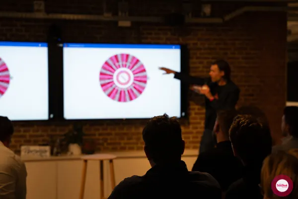 Live at SaaStock Local Amsterdam Q2 2024 — wider, low-light view of the audience watching the prize wheel spin on the dual screens at the front of the room