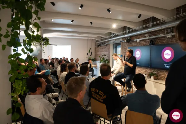 Live at SaaStock Local Amsterdam Q2 2024 — overview of the audience watching the panel through trailing pothos plants in the foreground, with the SaaStock Local Amsterdam screens lit at the front