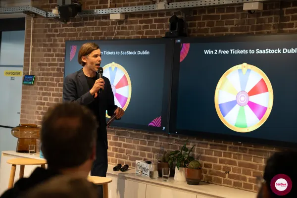 Live at SaaStock Local Amsterdam Q2 2024 — Ferdinand Goetzen at the mic in front of a 'Win 2 Free Tickets to SaaStock Dublin' slide showing a colourful prize wheel