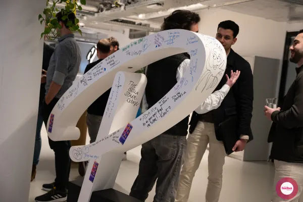 Live at SaaStock Local Amsterdam Q2 2024 — attendees signing a large white SaaStock 'R' / R-shaped sculpture covered in handwritten notes and messages from previous SaaStock events