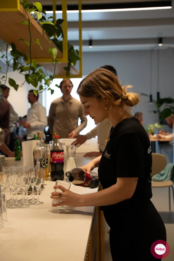 Live at SaaStock Local Amsterdam Q2 2024 — a server pours drinks at the bar at Personio's Amsterdam office, prosecco flutes lined up beside Coca-Cola and Royal Club mixers