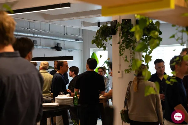 Live at SaaStock Local Amsterdam Q2 2024 — Amsterdam SaaS founders mingling in clusters at Personio's office before the talks begin, framed by hanging pothos plants