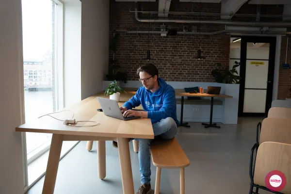 Before doors at SaaStock Local Amsterdam Q2 2024 — Joran Hofman in a blue Reditus hoodie working on a MacBook at a long wooden table beside Personio's Amsterdam office windows, with the canal view outside and a 'DAM SQUARE 202' meeting-room sign behind