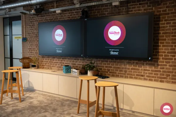Before doors at SaaStock Local Amsterdam Q2 2024 — the two dual screens at the front of Personio's event floor showing the 'SaaStock Local Amsterdam — Brought to you by Personio' opening slide before attendees arrived