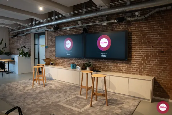 Before doors at SaaStock Local Amsterdam Q2 2024 — the SaaStock Local Amsterdam screens up at the front of Personio's event floor with three empty wooden stools positioned on a grey rug where the panelists would sit, Personio swag laid out on the cabinet below