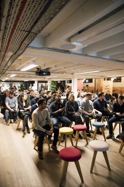Live at SaaStock Local London — packed audience seated on chairs and colorful red/yellow/blue stools, framed from a low angle