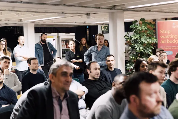 Live at SaaStock Local London — packed audience attentive to the talk, mixed standing and seated, a coral marketing sponsor pull-up banner reading 'Stop wasting ad budget' visible to the right under hanging plants