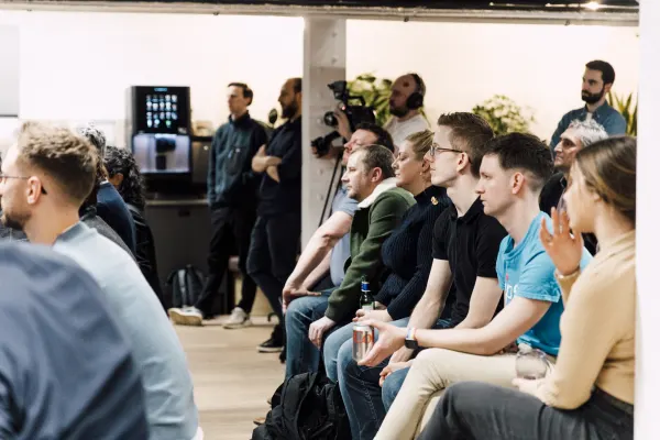 Live at SaaStock Local London — seated audience watching the talk with a camera operator on a tripod recording from the back of the room, coffee machine and crew silhouetted behind