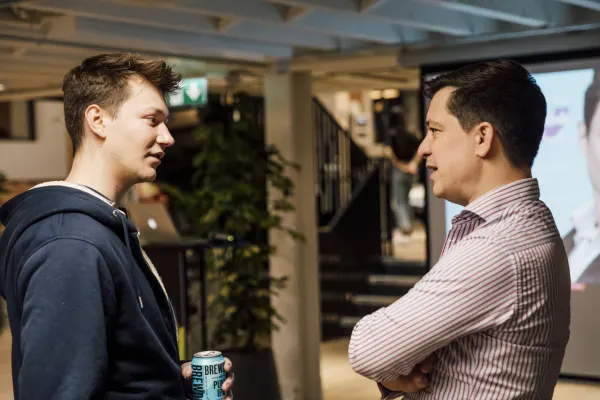 Live at SaaStock Local London — two attendees facing each other in conversation, one in a dark hoodie holding a BrewDog Punk IPA can, the other in a pink striped shirt with arms crossed, projection screen and staircase visible behind