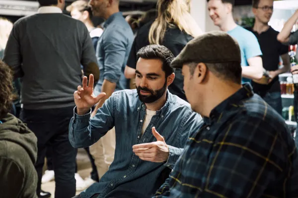 Live at SaaStock Local London — two founders in close conversation: bearded man in a denim shirt gesturing with both hands, the other in a grey flat cap listening, packed reception in the background
