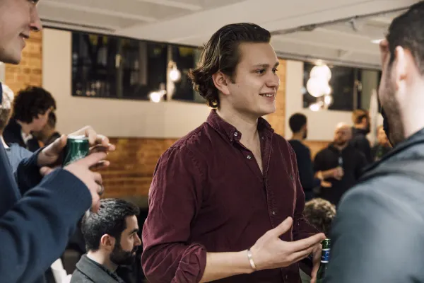 Live at SaaStock Local London — attendee in a maroon corduroy shirt mid-conversation in the reception area, drinks in hand, other founders in a cluster around him