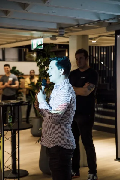 Live at SaaStock Local London — speaker in a blue-and-white striped shirt at the microphone, a second person in a dark t-shirt standing behind him near a column of plants