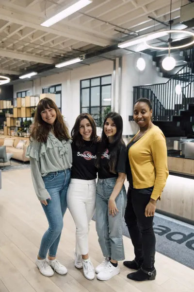 Live at SaaStock Local London — group portrait of four women from the SaaStock team and event crew, two in black SaaStock-branded t-shirts, smiling together in the bright modern lobby with the venue staircase behind them